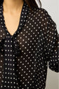 A person wearing a sheer black blouse with white polka dots and a tied bow at the collar. Only the upper torso and part of one arm are visible.