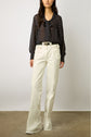 A woman stands against a plain wall wearing a black, long-sleeve, polka-dot blouse with a bow at the neck, tucked into high-waisted, wide-leg white jeans with frayed hems and a black belt. Her face is not visible.