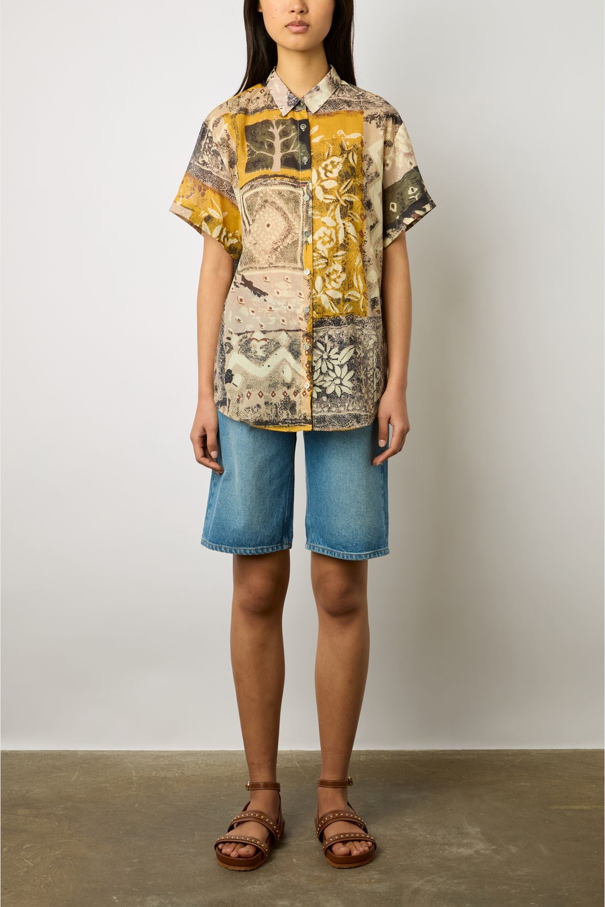 A person wearing a short-sleeve chemisette coton patterned button-up shirt with imprimé africain, knee-length denim shorts, and brown sandals stands against a plain light background. Their face is partially out of frame.