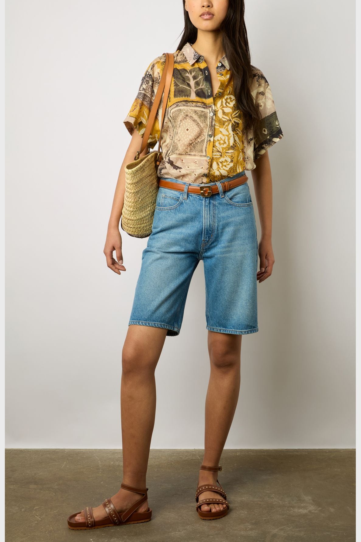 Woman wearing a short-sleeve chemisette coton shirt with an imprimé africain, blue knee-length denim shorts, brown sandals, a brown belt, and carrying a woven straw shoulder bag, standing against a plain background.
