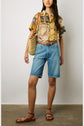 Woman wearing a short-sleeve chemisette coton shirt with an imprimé africain, blue knee-length denim shorts, brown sandals, a brown belt, and carrying a woven straw shoulder bag, standing against a plain background.