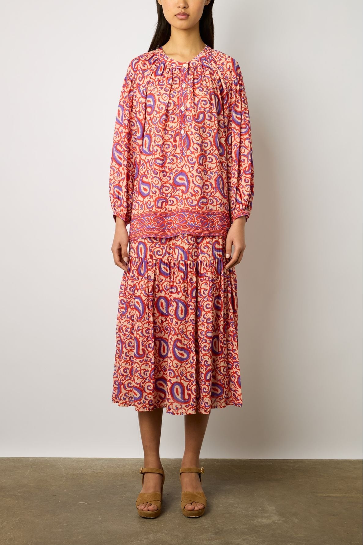 Woman wearing a long-sleeved, loose-fitting dress with a pink, red, and purple paisley imprimé ethnique, standing against a plain wall and brown heeled sandals—a striking look from the latest collection Studio.