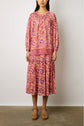 Woman wearing a long-sleeved, loose-fitting dress with a pink, red, and purple paisley imprimé ethnique, standing against a plain wall and brown heeled sandals—a striking look from the latest collection Studio.