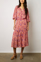 A woman stands wearing a colorful, patterned, long-sleeve midi dress with a tiered skirt from the collection Studio, paired with tan platform sandals against a plain wall.