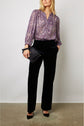 A woman wearing a purple patterned blouse tucked into black pants stands holding a black clutch bag, with one hand in her pocket, against a plain background.
