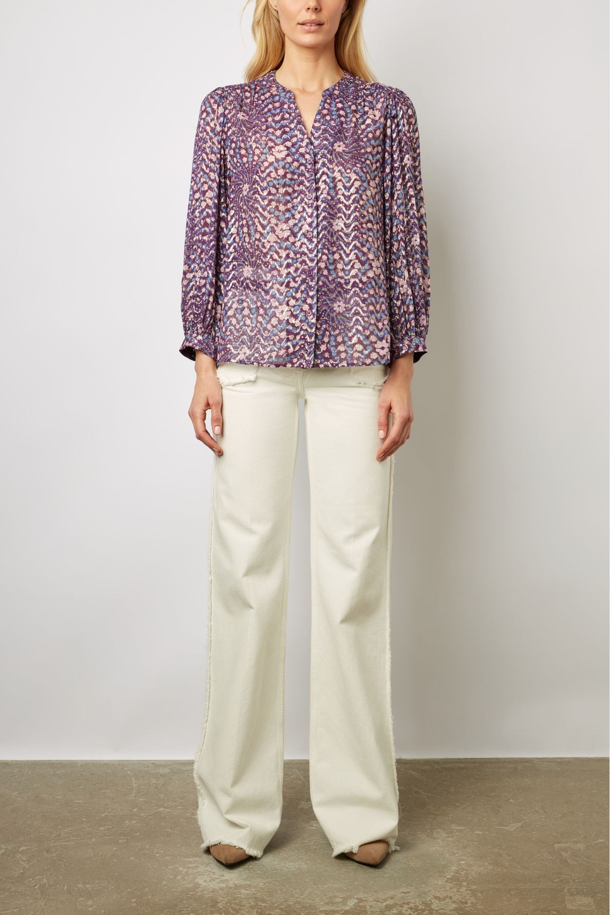 A woman wearing a purple patterned blouse with white buttons and wide-legged white pants stands against a plain background. Her face is partially out of frame.