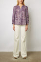 A woman wearing a purple patterned blouse with white buttons and wide-legged white pants stands against a plain background. Her face is partially out of frame.