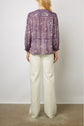 A woman stands facing away, wearing a patterned purple blouse with long sleeves and loose white pants, on a concrete floor with a white wall background.