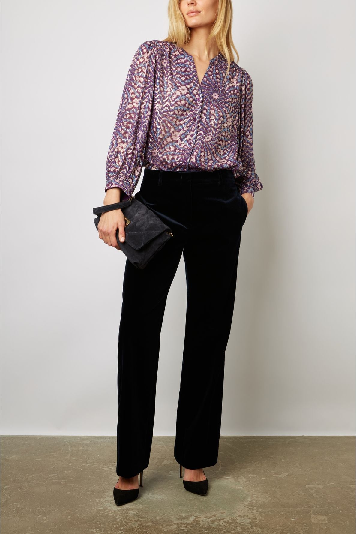 Woman wearing a purple patterned blouse, black wide-leg pants, and black heels holds a black clutch. She stands against a plain light-colored wall, looking slightly to the side.