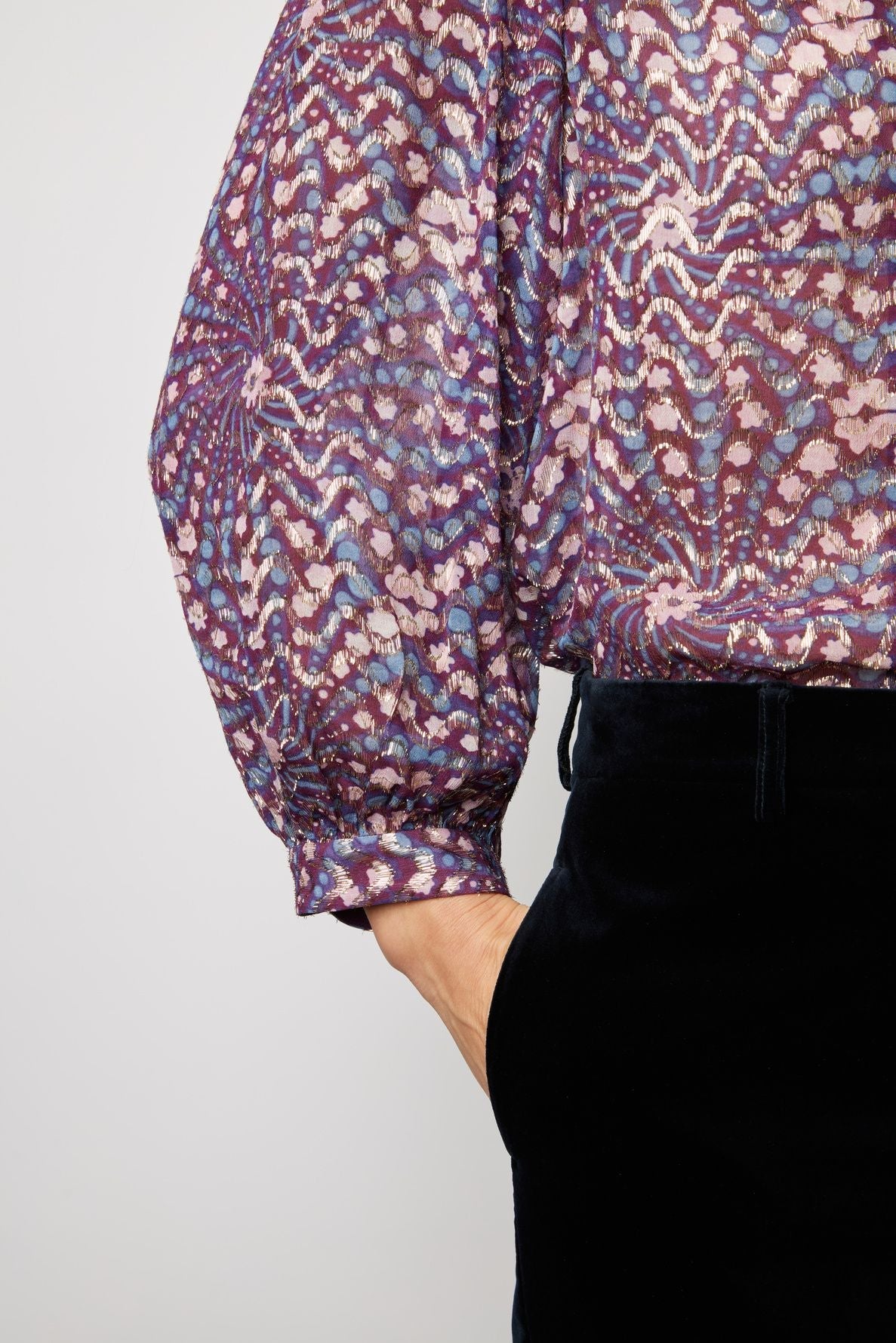 A person wearing a colorful, patterned long-sleeve blouse with a purple and pink design, and black pants, has their right hand tucked into their pocket. The background is plain and light.