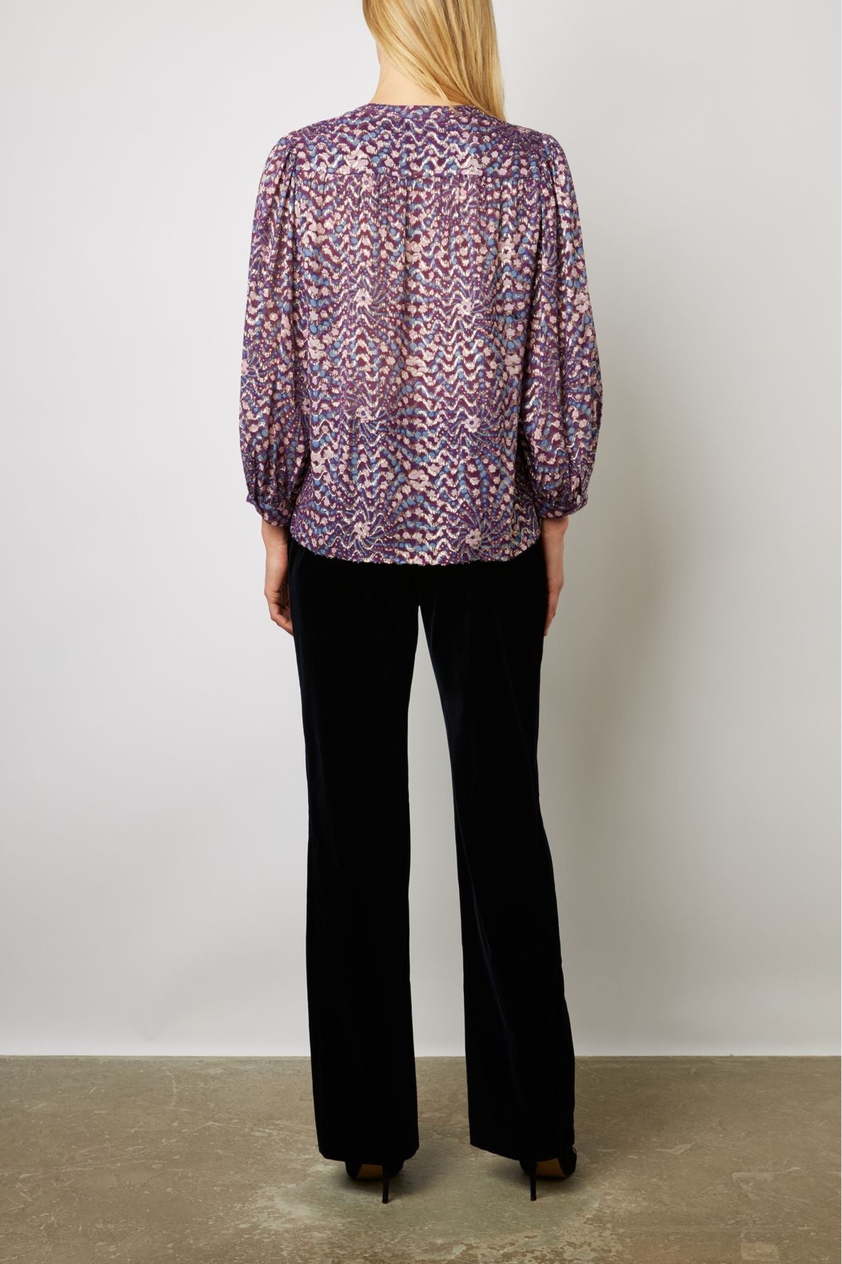 A woman stands facing away, wearing a purple patterned blouse with three-quarter sleeves and loose black pants. She has straight blonde hair, and her arms are relaxed by her sides. The background is plain and light-colored.