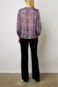 A woman stands facing away, wearing a purple patterned blouse with three-quarter sleeves and loose black pants. She has straight blonde hair, and her arms are relaxed by her sides. The background is plain and light-colored.