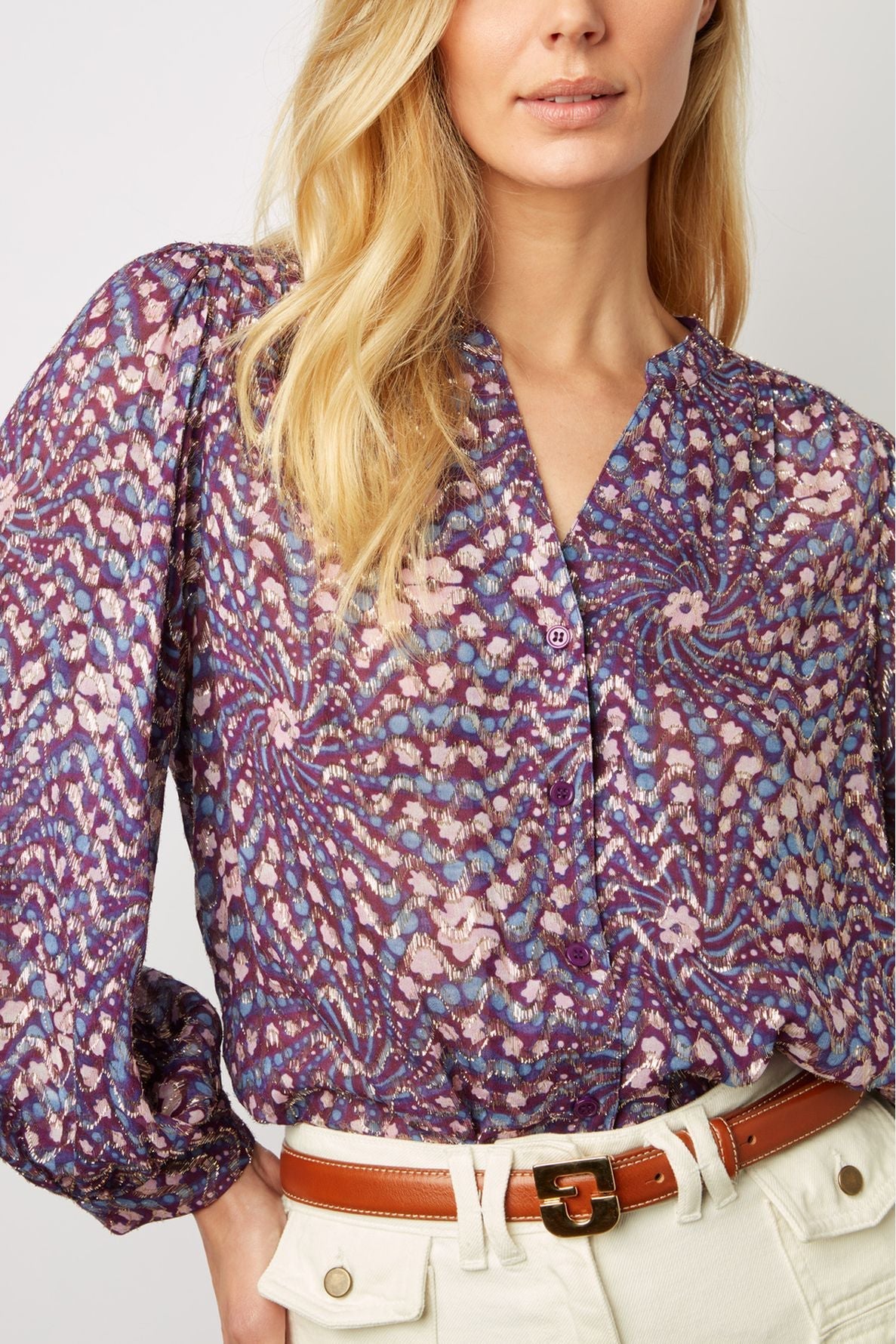 Woman wearing a purple patterned blouse with long sleeves and cream high-waisted pants, accessorized with a tan belt. The image is cropped at the shoulders and waist.
