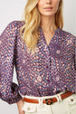 Woman wearing a purple patterned blouse with long sleeves and cream high-waisted pants, accessorized with a tan belt. The image is cropped at the shoulders and waist.