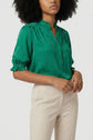 A woman with curly hair wears a bright green, patterned blouse with short sleeves and beige pants, standing against a plain light background.