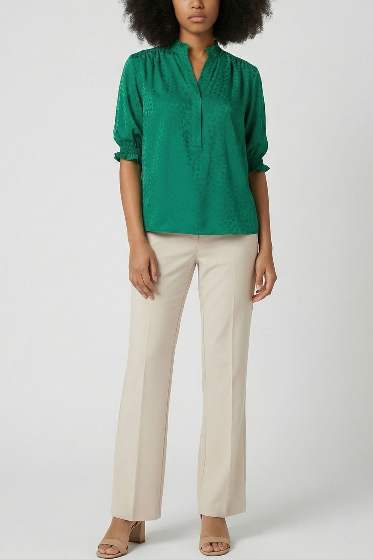 A person wearing a green long-sleeve blouse, beige pants, and beige heeled sandals stands against a plain light background. The outfit is neat and semi-formal.