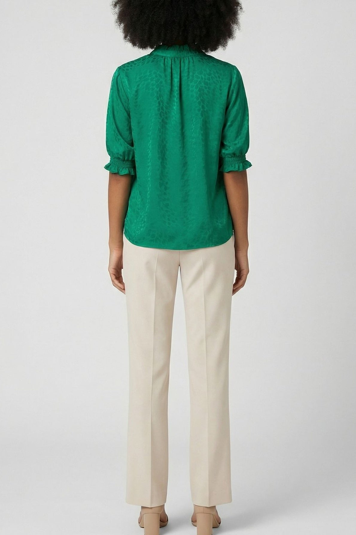 A person with curly hair is standing facing away, wearing a green blouse with three-quarter sleeves and beige trousers with heels, against a plain light background.