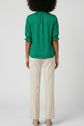 A person with curly hair is standing facing away, wearing a green blouse with three-quarter sleeves and beige trousers with heels, against a plain light background.
