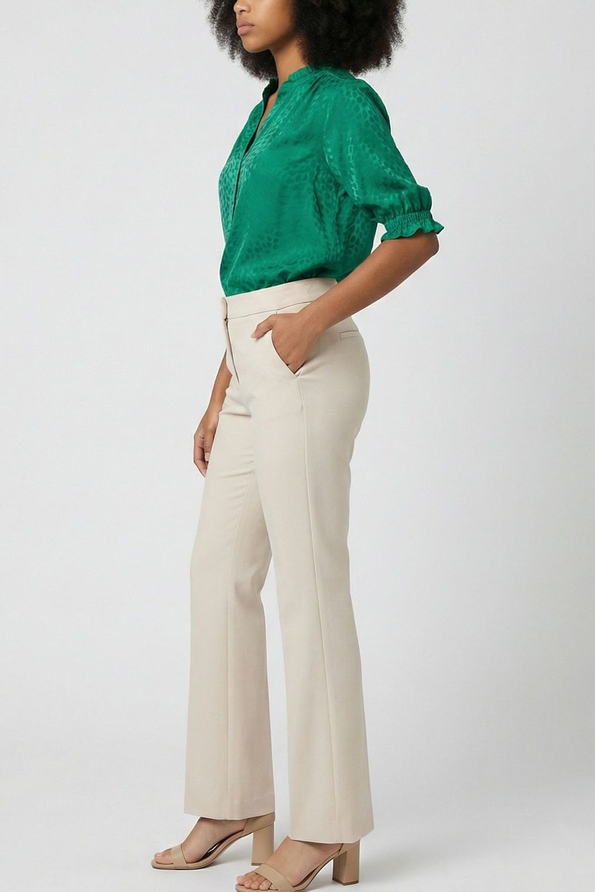 Woman standing sideways, wearing a green blouse with elbow-length sleeves, beige high-waisted trousers, and beige heeled sandals. Her left hand is in her pocket, and she has curly shoulder-length hair. The background is plain.
