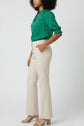 Woman standing sideways, wearing a green blouse with elbow-length sleeves, beige high-waisted trousers, and beige heeled sandals. Her left hand is in her pocket, and she has curly shoulder-length hair. The background is plain.