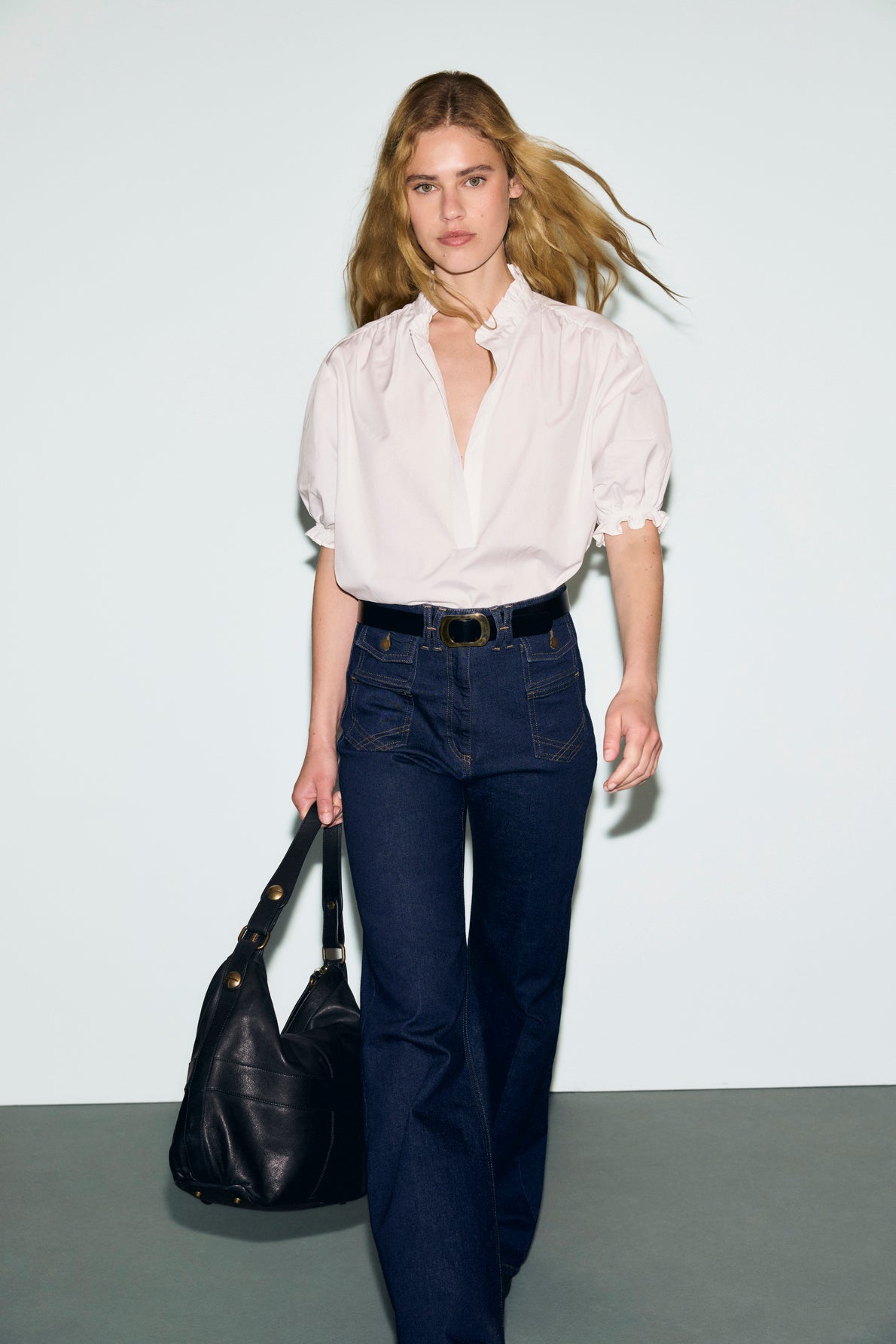 A woman with long blonde hair walks forward wearing a white blouse coton with puff sleeves, dark blue jeans, and a black belt, holding a large black bag. She stands against a plain, light-colored background.