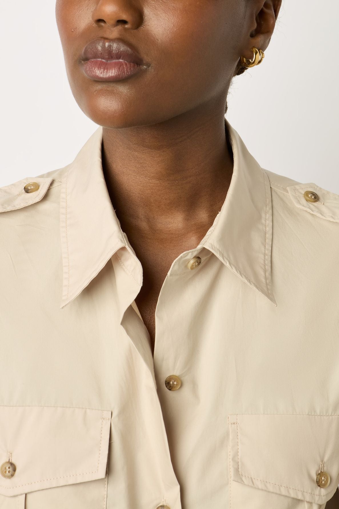 A woman wearing a beige button-up shirt with two front pockets and gold earrings, shown from shoulders to mouth.