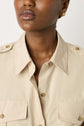 A woman wearing a beige button-up shirt with two front pockets and gold earrings, shown from shoulders to mouth.