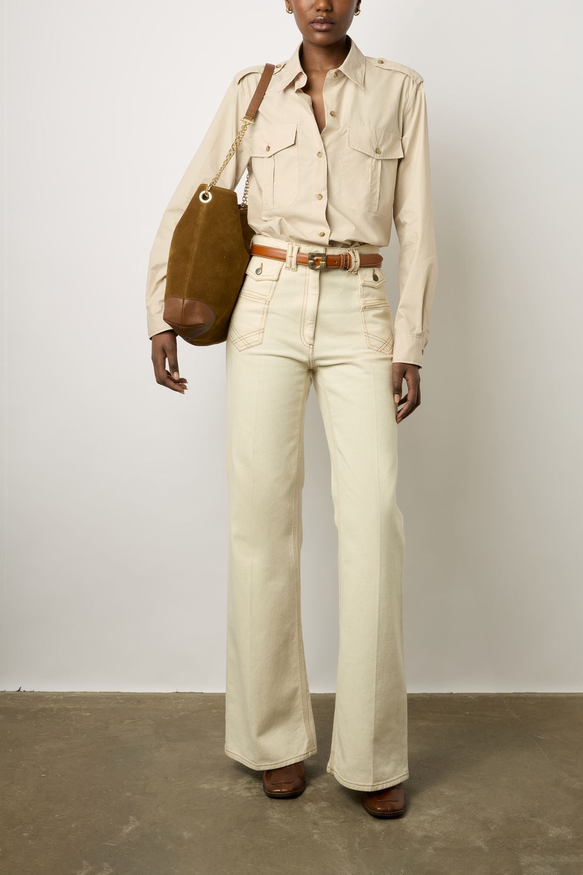 A person wearing a beige button-up shirt, high-waisted cream pants, brown belt, and brown shoes stands holding a brown shoulder bag against a plain white wall.