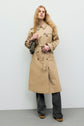 A woman with long blonde hair wears a trench long camel coton, gray patterned pants, and platform shoes. She holds a small gray bag and stands against a plain light background, showcasing the perfect manteau mi-saison look.
