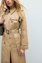 A woman with long blonde hair wears a trench long camel en coton, featuring a belt and double-breasted buttons, while holding a green purse with a chain strap. Her face is partially out of frame against a plain light background, highlighting this parfait manteau mi-saison.