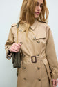A person wearing a trench long camel en coton, styled as a classic manteau mi-saison, and carrying a green shoulder bag with a gold chain strap stands against a plain light background.