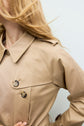 A person with long, light brown hair wears a trench camel coton beige trench coat with two visible buttons, posing with one hand on their hip against a plain light background.