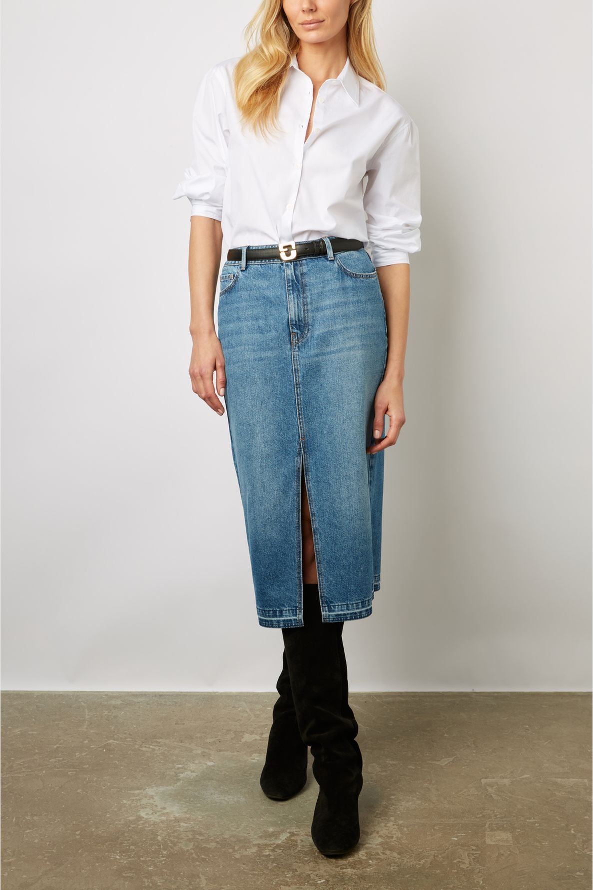 Woman wearing a white button-down shirt tucked into a knee-length denim skirt with a front slit, black belt, and black knee-high boots, standing against a plain background.