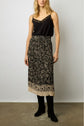 A woman wearing a sleeveless black top with lace trim, a black and beige motif floral jupe midi plissée crafted from crêpe georgette, and black knee-high boots stands against a plain white wall.
