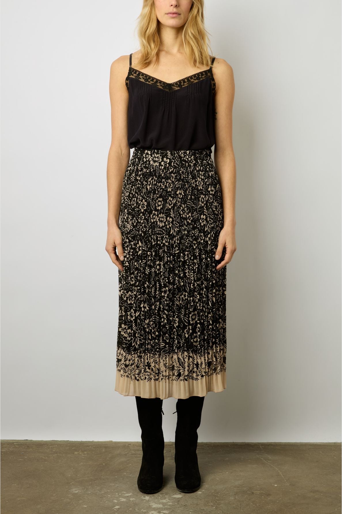 A woman wearing a black sleeveless top and a pleated, black and beige patterned jupe midi plissée stands facing forward against a plain light-colored wall, with her hands by her sides and black boots on.