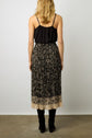 A woman with blonde hair stands facing away, wearing a black sleeveless top and a jupe midi plissée in motif floral crêpe georgette, paired with tall black boots, against a plain light background.