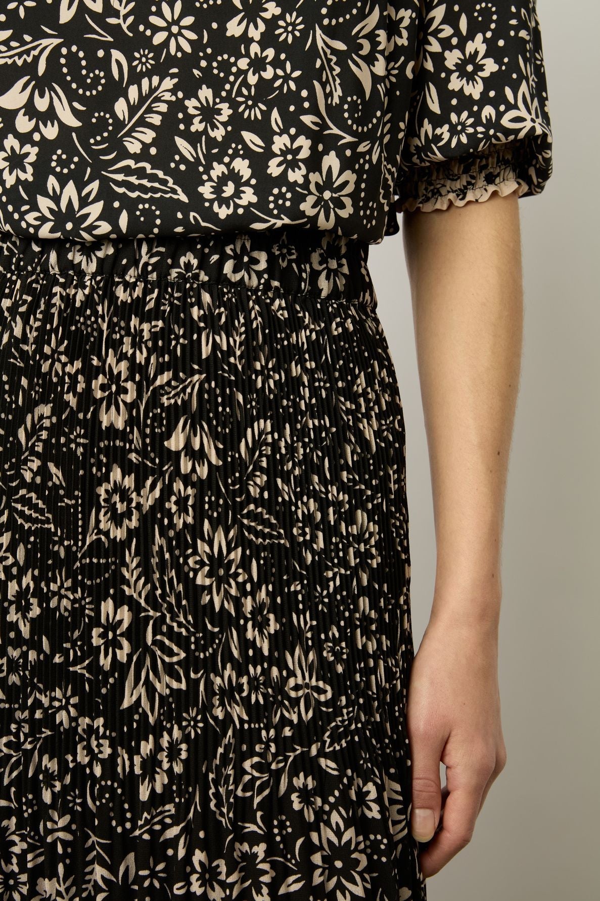 A person wearing a black crêpe georgette dress with a beige motif floral pattern stands against a plain background. Only the side of the torso, arm, and hand are visible.