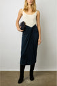 A woman wears a white sleeveless top, a navy knotted midi skirt, and black boots. She holds a small dark clutch and stands against a plain white wall on a concrete floor.
