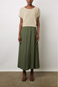 A person stands against a white wall wearing a beige short-sleeve top, an olive green jupe longue satinée, and black heeled sandals. Their arms are relaxed by their sides.