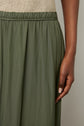 A close-up of a person wearing an olive green, flowy jupe plissée with an elastic waistband, reflecting the kaki tendance. Their arm hangs by their side, and their hand is relaxed, showing neatly manicured nails. The top is a light beige, textured fabric.