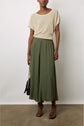A person wearing a short-sleeve beige top, loose olive green wide-leg pants inspired by the kaki tendance, black strappy sandals, and holding a black bag, stands against a plain white wall on a concrete floor.