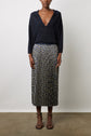 A woman stands against a plain background wearing a deep navy V-neck top with long sleeves and a jupe plissée longueur midi featuring striking motifs géométriques in black, white, and yellow. She completes the look with black strappy sandals.