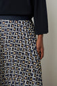 A person wearing a dark long-sleeve top and a jupe plissée with motifs géométriques in navy, white, and tan stands against a neutral background.