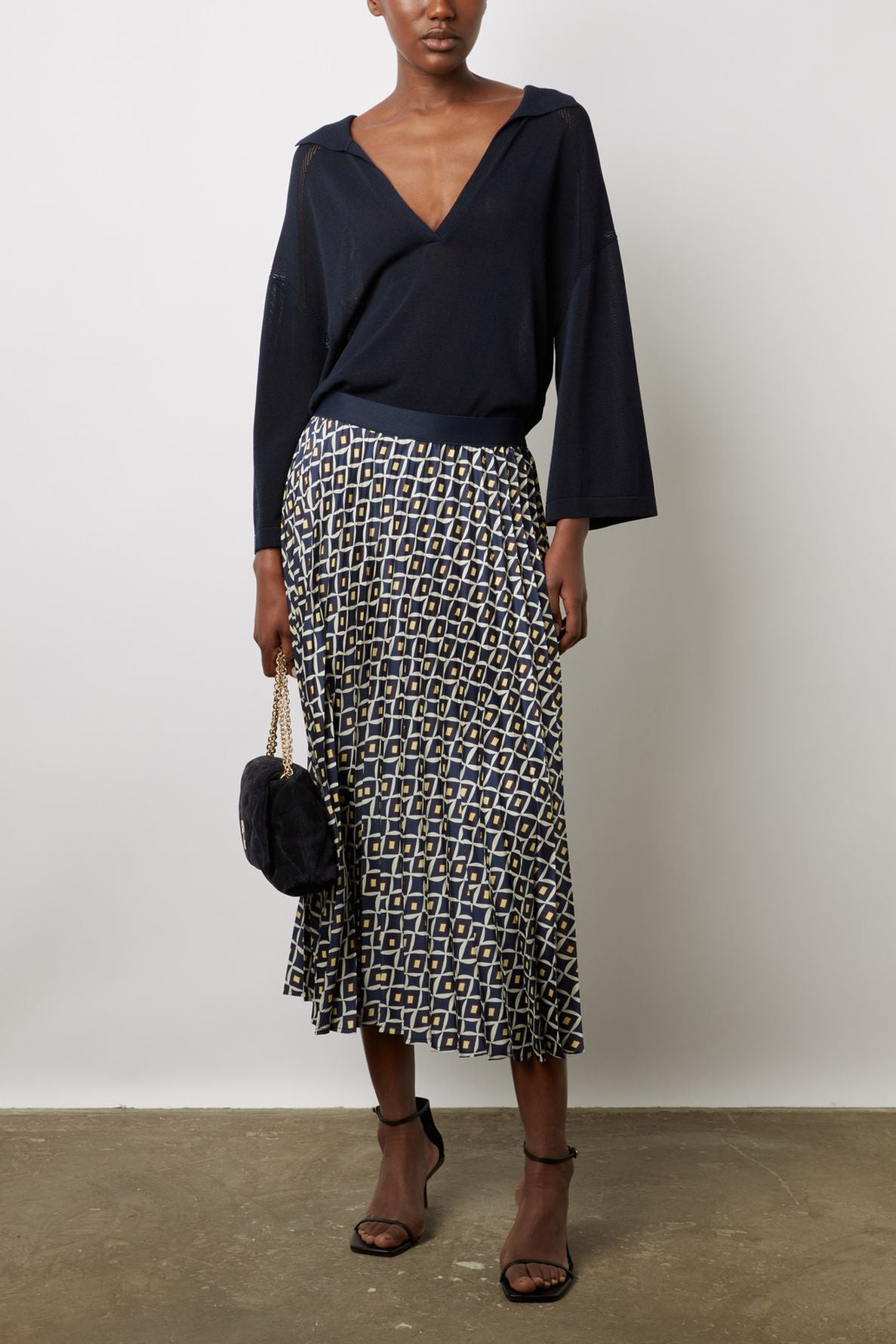 A person wearing a navy V-neck sweater, a jupe plissée longueur midi with motifs géométriques, black strappy heels, and holding a black handbag with a gold chain stands against a plain white wall.