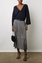 A person wearing a navy V-neck sweater, a jupe plissée longueur midi with motifs géométriques, black strappy heels, and holding a black handbag with a gold chain stands against a plain white wall.