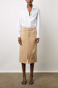 A person stands against a plain background wearing a white button-up shirt, a tan knee-length skirt with a front slit, and black strappy heels. Their face is not fully visible.