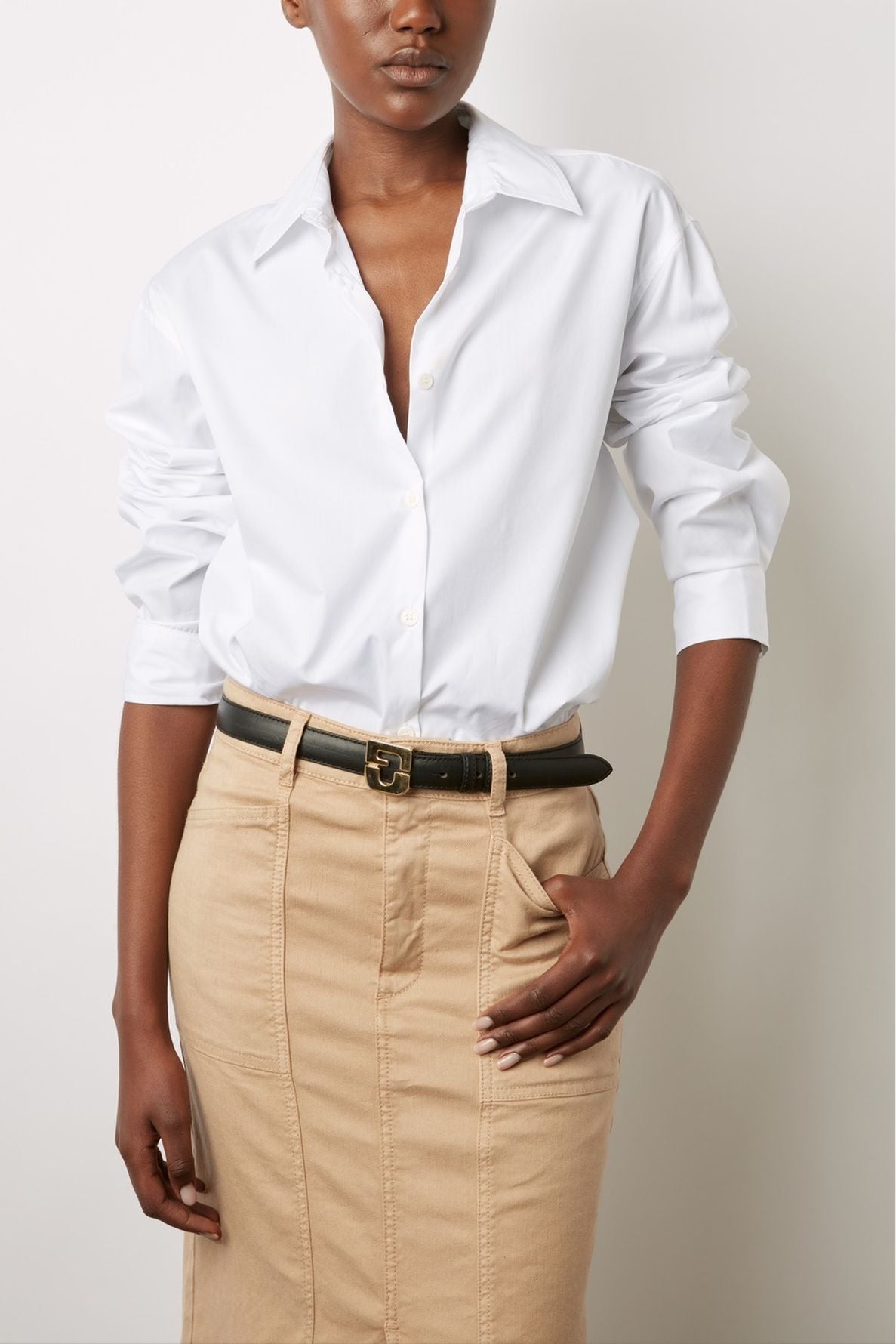 A person wearing a white button-up shirt tucked into a high-waisted beige skirt, accessorized with a black belt, stands against a plain background with one hand in a pocket.