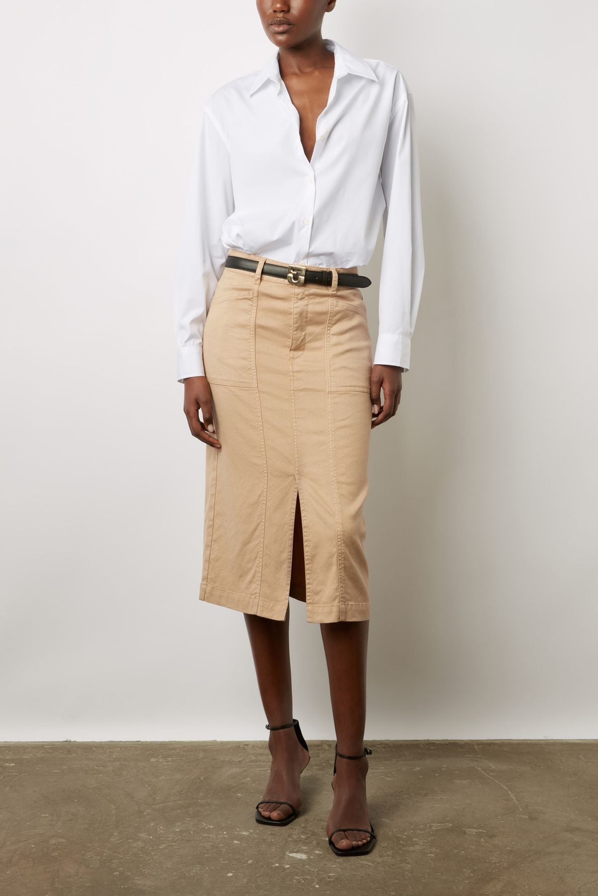 A person wears a white button-down shirt, tan knee-length skirt with a front slit, black belt, and black strappy heels, standing against a plain white wall.