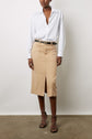 A person wears a white button-down shirt, tan knee-length skirt with a front slit, black belt, and black strappy heels, standing against a plain white wall.