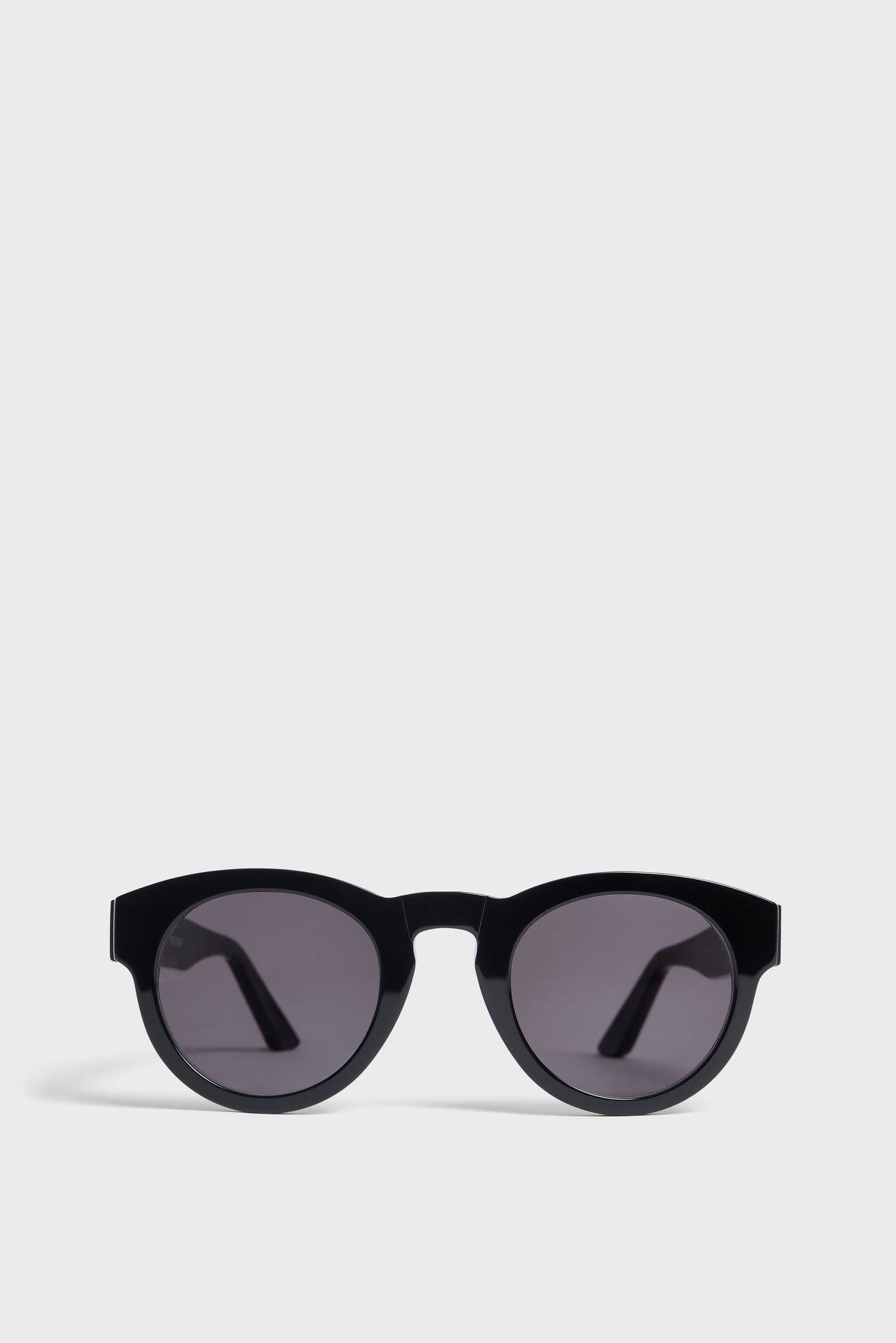 Black round lunettes de soleil with dark lenses are displayed against a plain light gray background. Offering excellent protection UV, the sunglasses feature thick frames and slightly angled arms, and they are oriented with the lenses facing forward.